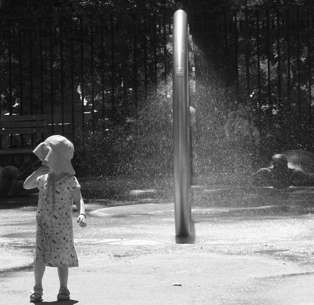 A little girl contemplates running through the sprinkler alone. 